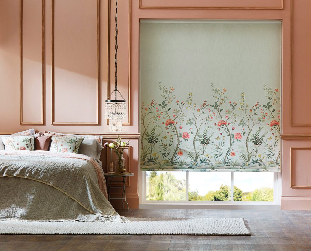 Image shows a bedroom interior with pink panelled walls and a small chandelier over the bedside table. The window is covered with our DIY roller blind in a fabric called Chinoiserie in a colour called Duck Egg. The design features whimsical flowers and leaves that seem to grow from the bottom of the blind. 