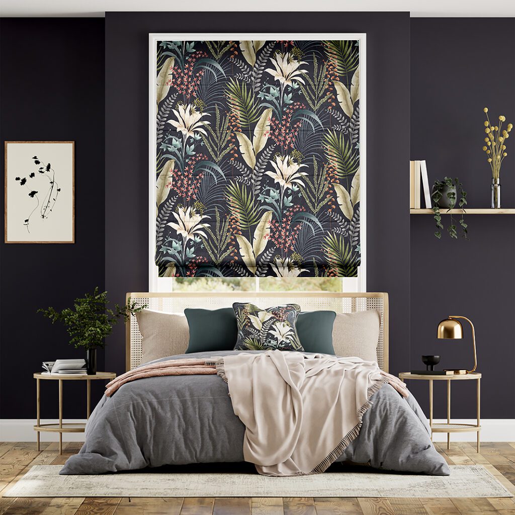 Image shows a roman blind with blockout lining in a bedroom interior. There is a tropical print on the fabric of the blind against a dark painted wall with a bed with grey and cream linens. 