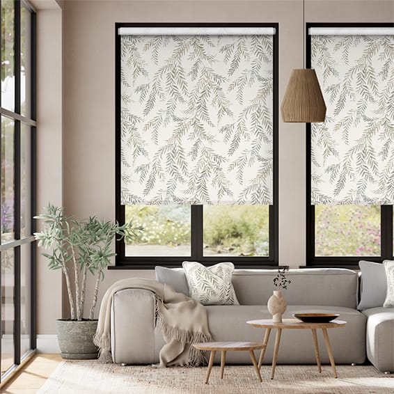 Image shows a modern sitting room with a low contemporary sofa and Scandinavian coffee tables. The windows are covered with DIY roller blinds from Tuiss Blinds Online in a fabric called Dappled Ferns, which has an organic pattern of fern leaves on a neutral background. 