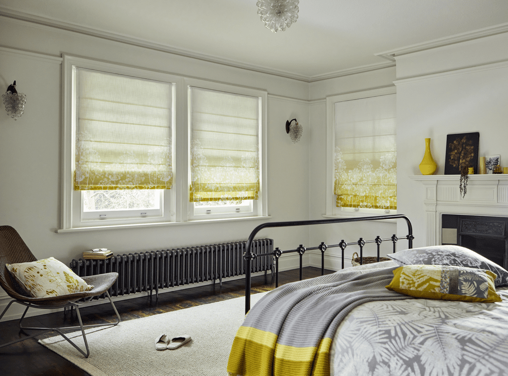 Image shows a bedroom interior with a colour palette of white, yellow and grey. The windows are covered with roman blinds from the Clarissa Hulse collection from Tuiss. They show what thermal interlining looks like on a designer blind. 