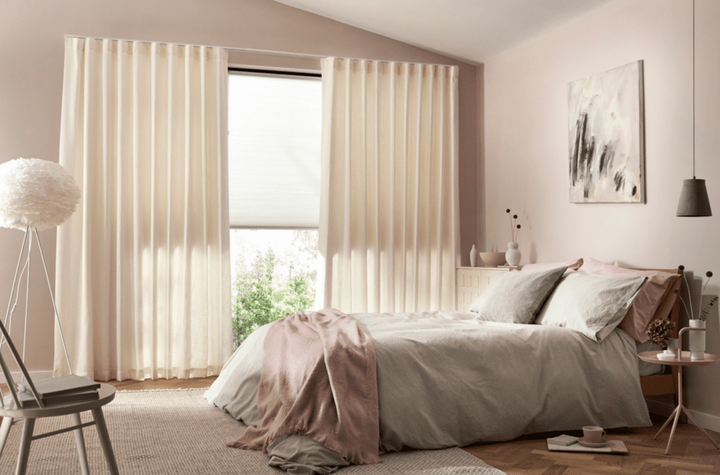 Image shows a bedroom interior with warm neutral tones and soft pink highlights on the bed. The window is covered with a honeycomb blind and an s-fold curtain with thermal interlining.