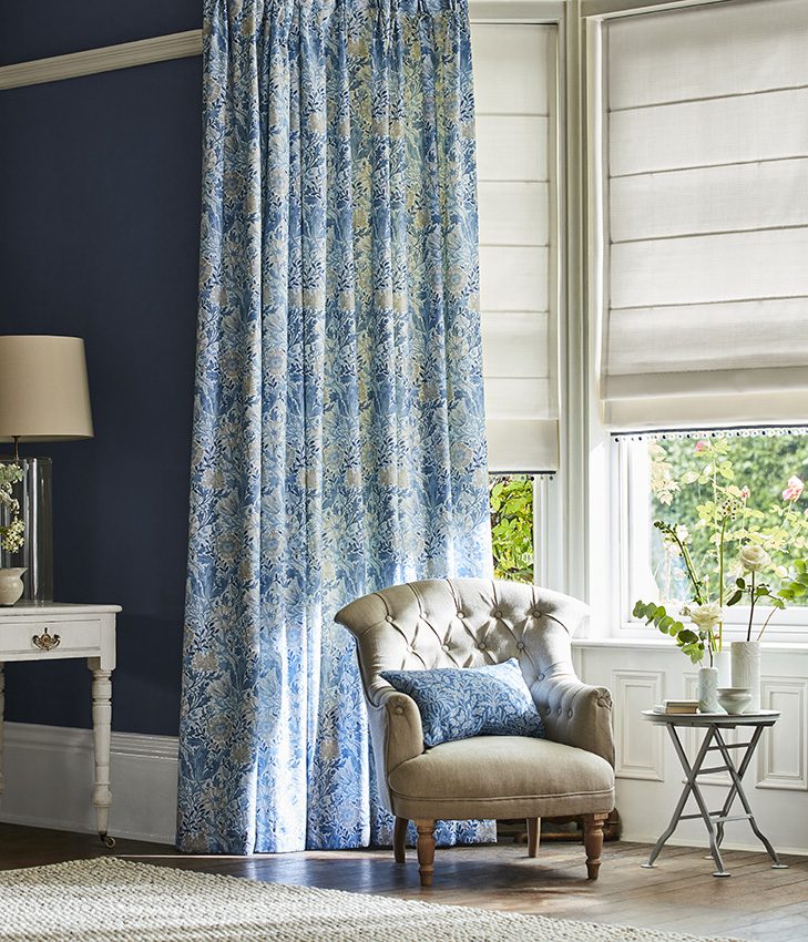 William Morris curtain in a blue design is shown layered over a roman blinds with a French provincial chair in front of the window. 