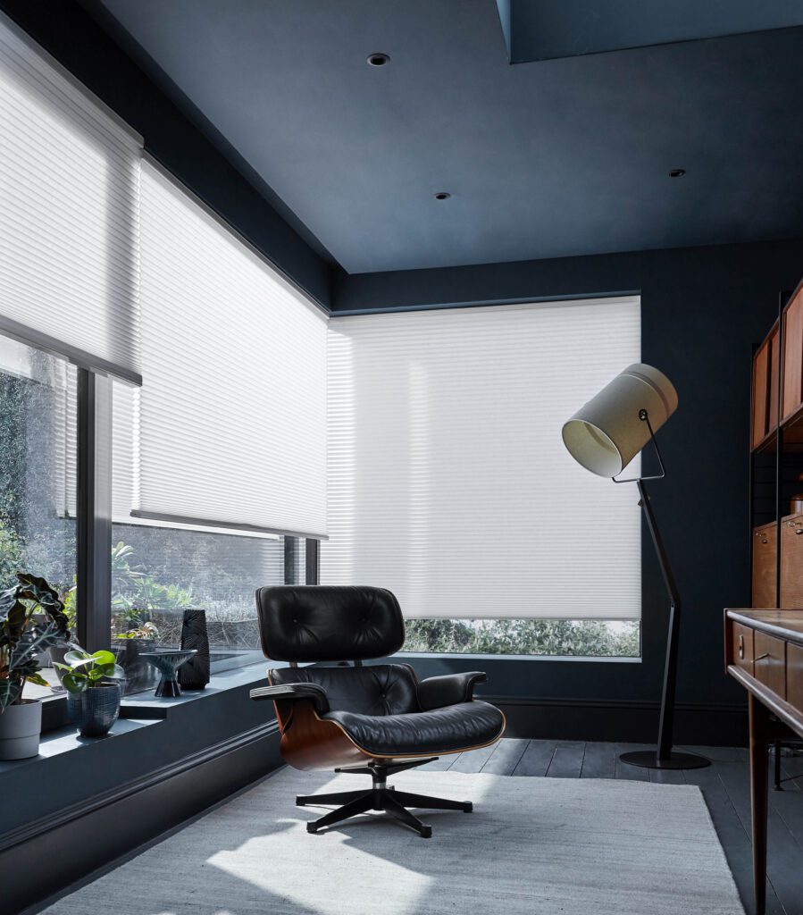 The honeycomb blinds in this image work well as blinds to keep you cool in summer.  The image shows an Eames lounge chair in black leather. The walls and ceiling are painted black, with white light filtering honeycomb blinds to save energy. 