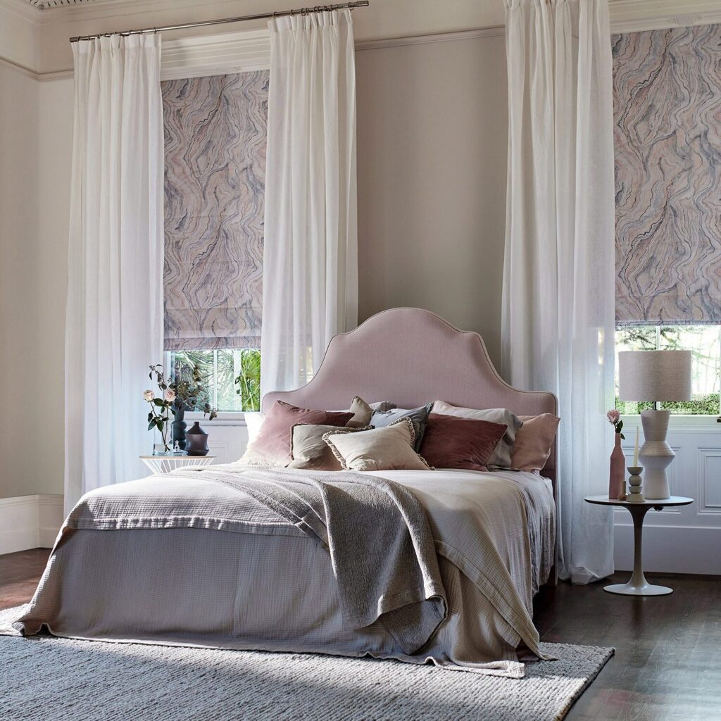 Image shows a bedroom interior with a soft pink and white colour palette. There are pink marbled roman blinds layered with white sheer curtains. 