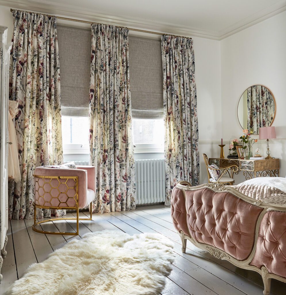 A french inspired interior with gilt framed bedhead with rose coloured tufted upholstery. The window are covered with linen roman blinds and beautifully patterned floral curtains. 