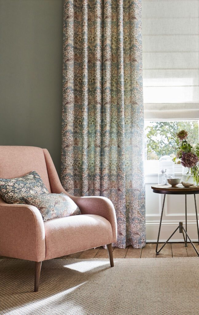 Sitting room interior with a pink armchair in front of a window. The window coverings are a white linen roman blind layered with William Morris curtains in the famous pattern 'Strawberry Thief'. 