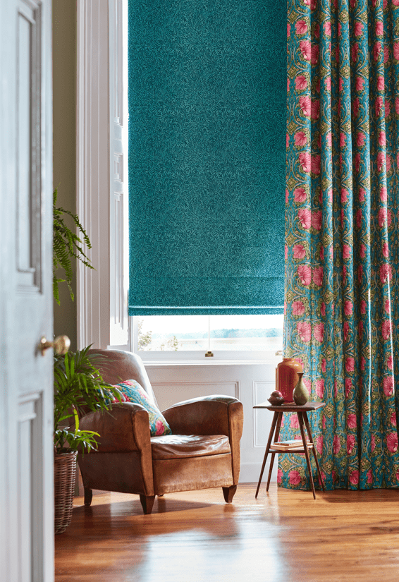 Sitting room with timber floorboards, leather armchair and William Morris window furnishings, demonstrating how to layer roman blinds and curtains. 